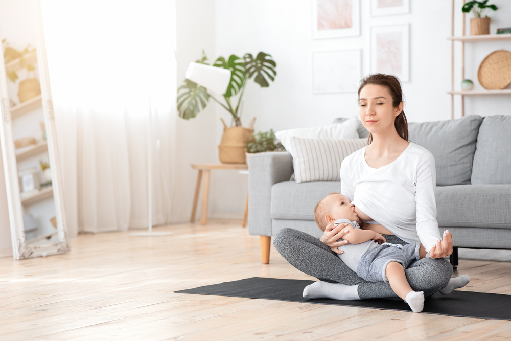 Relaxed Young Mother Meditating While Breastfeeding Her Baby At Home, Sitting In Lotus Position, Practicing Yoga, Doing Breathing Exercise, Copy Space
