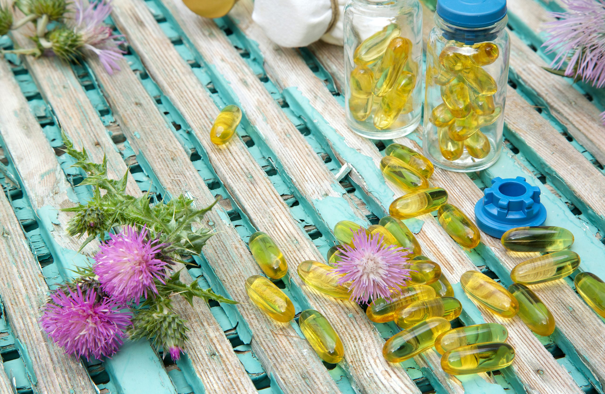 MILK THISTLE OIL IN SOFTGELS AND MILK THISTLE BLOSSOMS IN THE BACKGROUND